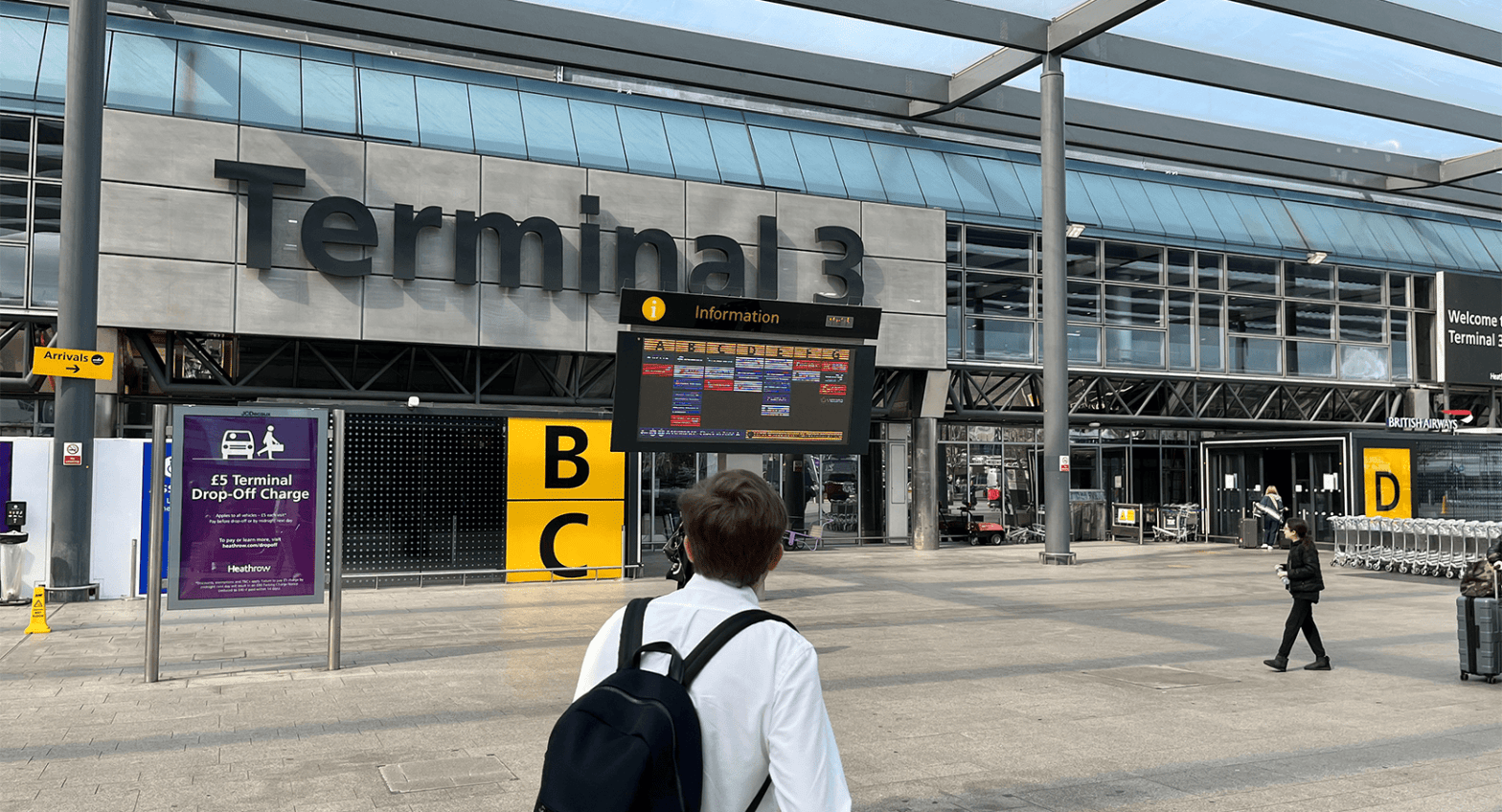 A man wearing a navy blue backpack and white shirt standing in front of Terminal 3 at London Heathrow Airport with B and C in bold yellow blocks.