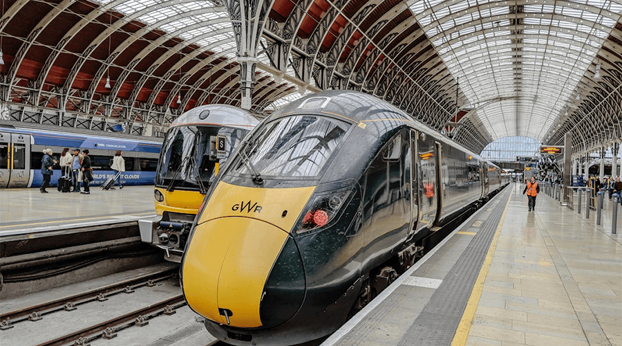 A green and yellow Great Western Railway train parked at a station with other trains and passengers in the background and train worker wearing an orange hi-vis vest.