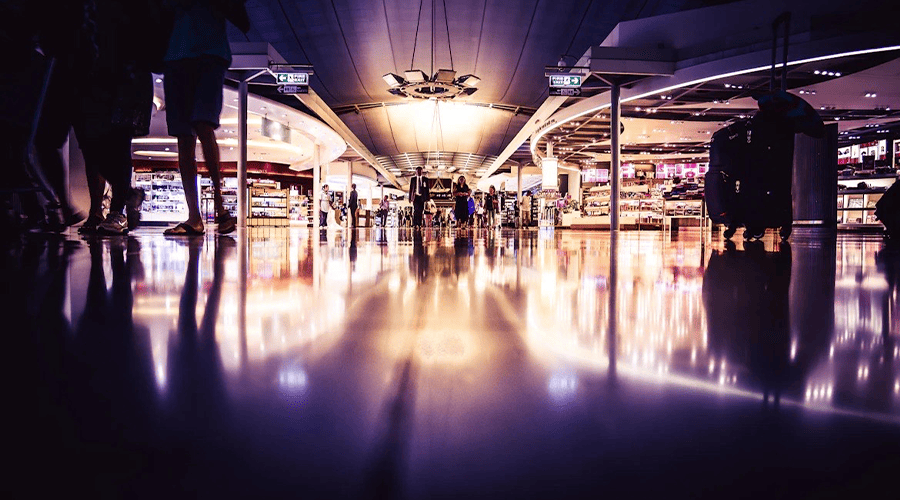 An airport duty free in dark lighting with passengers walking in the background and bright lights coming from the duty free kiosks.