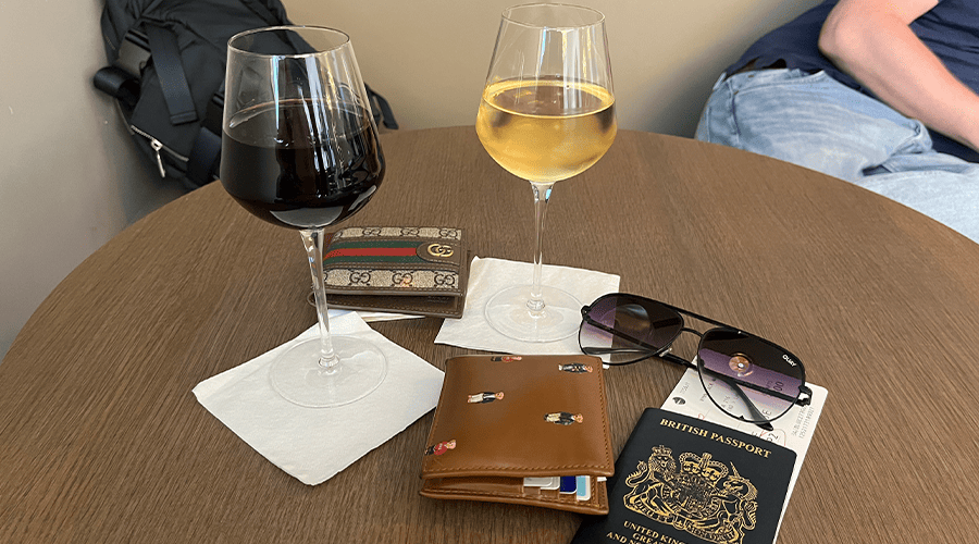 A glass of red wine and glass of white wine on a table on top of two white napkins, with a British passport, sunglasses, wallets and a navy backpack.
