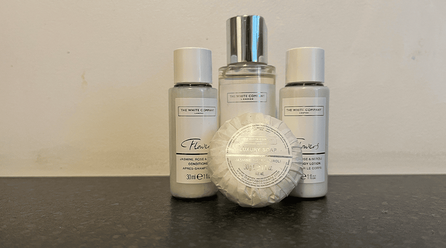 Four The White Company mini toiletries including a hand soap, shampoo, conditioner and body wash standing on a dark grey counter and off-white background.