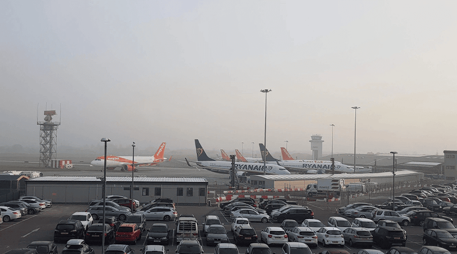 An airport car park with cars parked in front of Ryanair and EasyJet aircrafts at an airport terminal against a hazy grey sky.