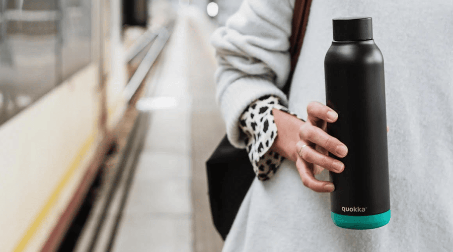 A female wearing a grey sweater standing next to a train on a train station platform holding a black and green reusable water bottle.