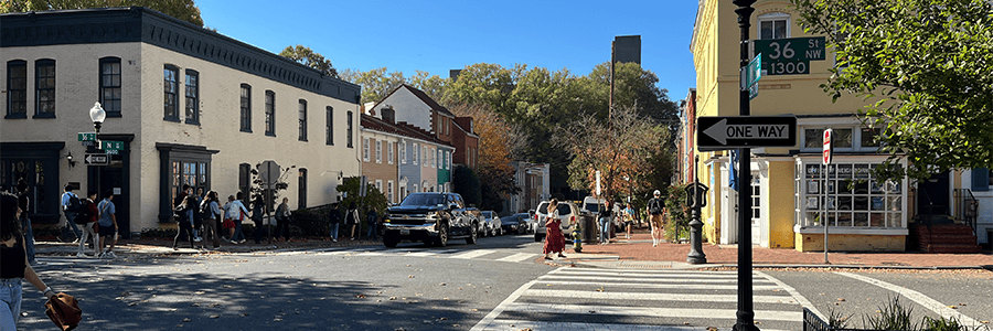 A picturesque street in Georgetown, Washington, D.C with a yellow building on the right against green trees and a clear blue sky with people crossing the streets.
