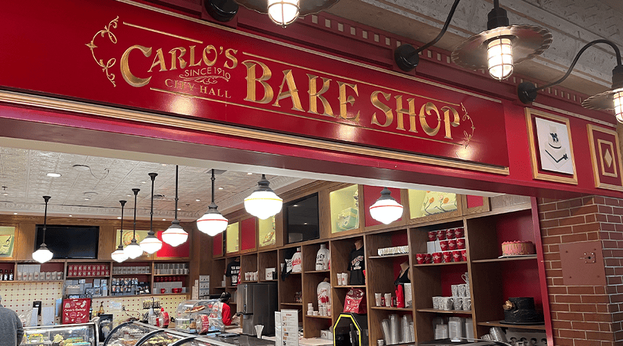 The red and gold sign for Carlo's Bakery at The Venetian Resort in Las Vegas against a brick wall and merchandise inside the bakery shop. 