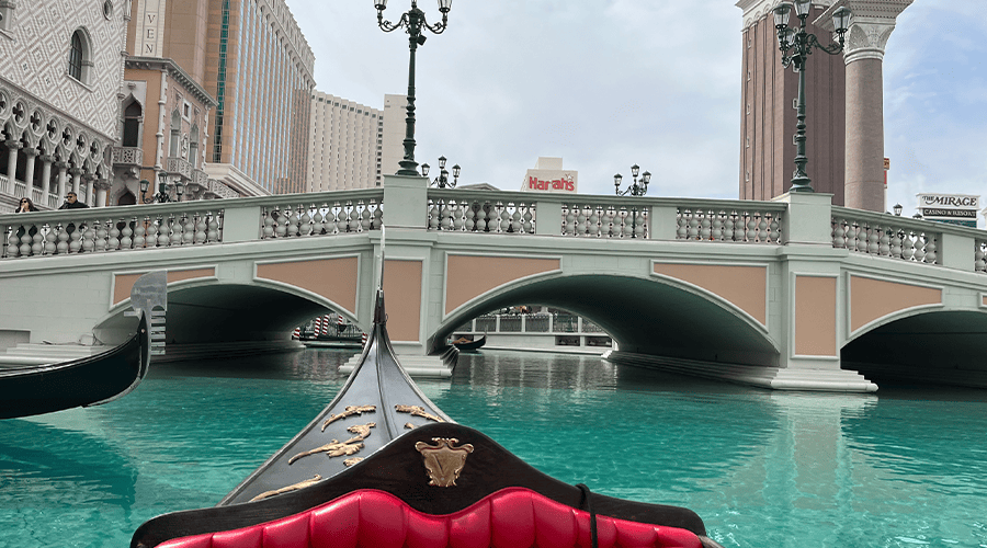 The outside Gondola ride at The Venetian Resort in Las Vegas with people standing on the bridge looking at the gondola and Harrah's in the distance against a cloudy sky. 