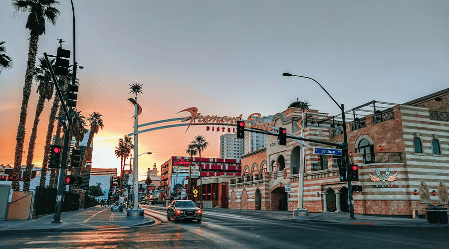 A sign for Fremont East District in Las Vegas next to palm trees and a car waiting at a red light and a sunset sky in the background.