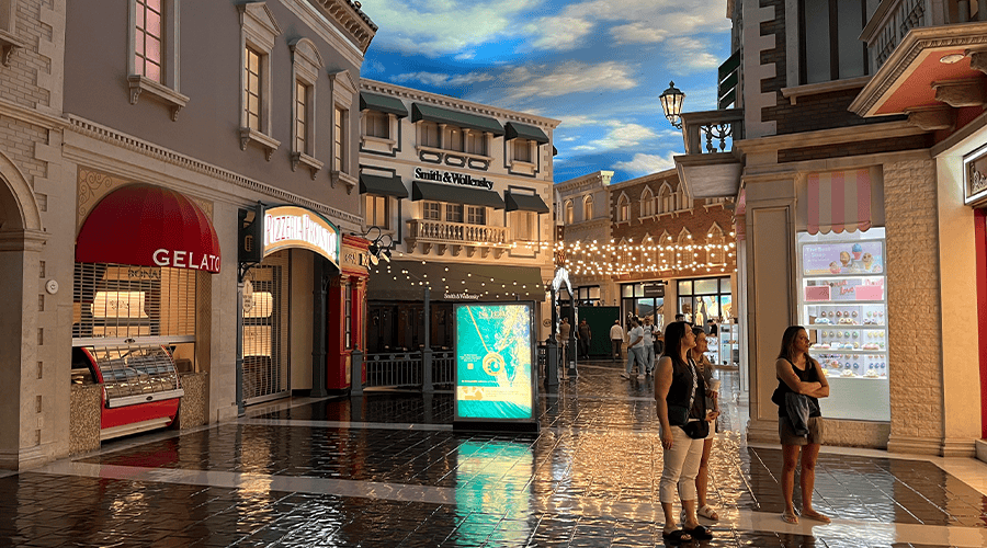 The Venetian Resort Grand Canal Shoppes in Las Vegas with people standing outside a shop opposite a Gelato stand against a blue cloudy sky and lights across the path. 