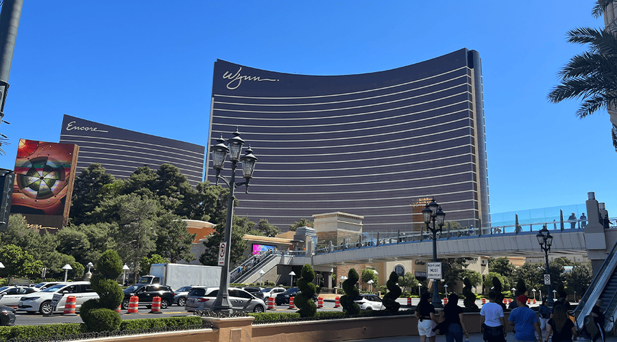 The Wynn Las Vegas hotel next to the Encore complex against a bright blue sky with cars on the road and people walking along the pathways and the bridge over the road against green trees and shrubbery. 