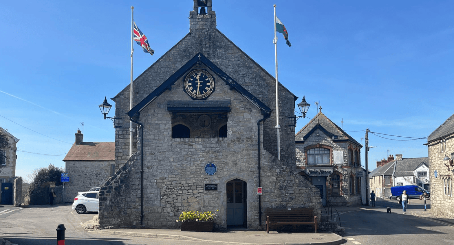 The front of Llantwit Major town hall with the UK and Welsh flags on either side against a bright blue sky and a woman walking a dog in the background.