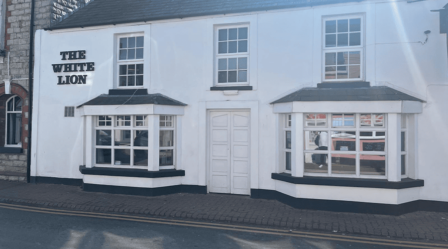 The outside of The White Lion Hotel in Llantwit Major with black lettering on the top right-side and a white entrance door, two big bay windows and three upstairs windows.