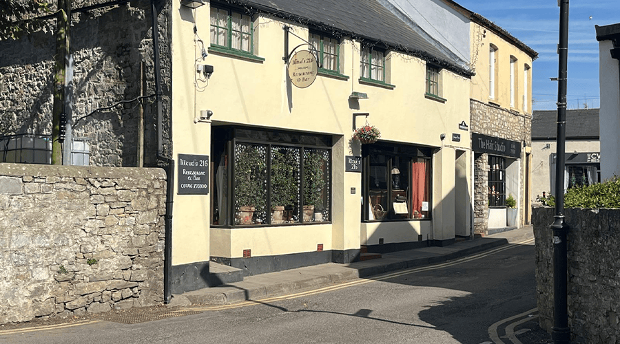 A yellow building with black and green window frames next to a brick wall which is the outside of Illtud’s 216 bistro in Llantwit Major with a hanging basket outside the entrance door.