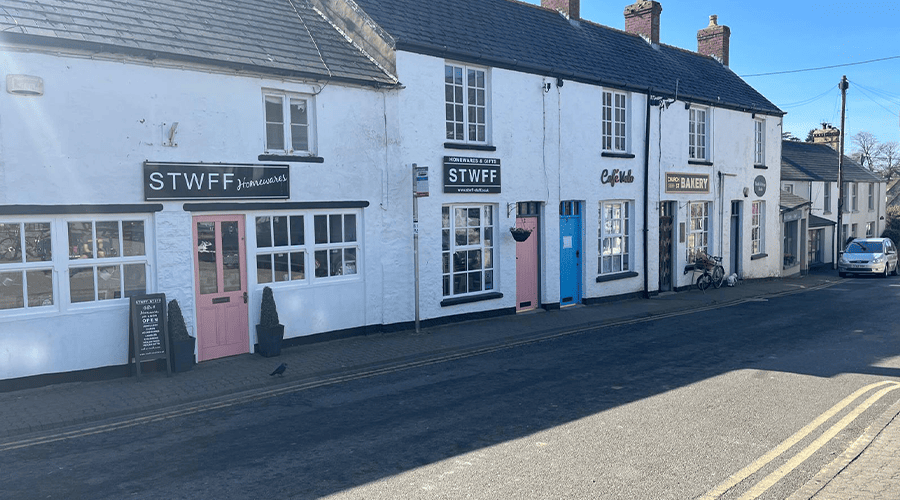 The outside of Stwff-Stuff in Llantwit Major which are white cottage buildings with pink and blue front doors alongside a concrete road and double yellow lines on the right-side of the road.