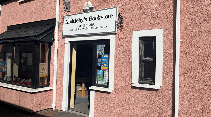 The outside of Nickelby's Bookstore in Llantwit Major which is painted in a shade of terracotta and the Nickleby's Bookstore sign is white in black writing and a window on either side of the entrance door.