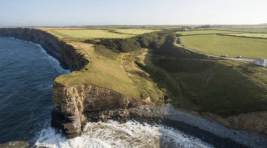An aerial view of the cliff tops at Llantwit Major with crashing eaves at the bottom against the rocky formations and cliffs with green fields and small white building on the right side.