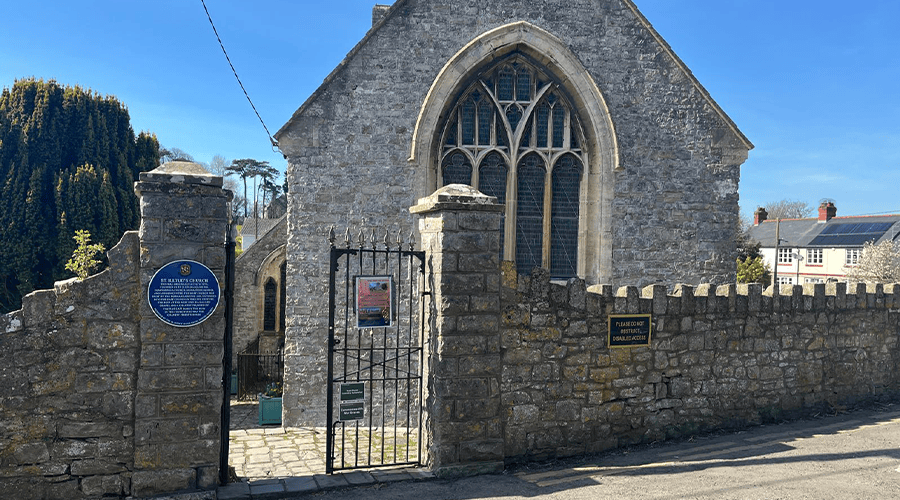 A front-facing view of St. Illtud’s Church in Llantwit Major with a blue plaque on the right side of the brick wall gate entrance against a blue sky and green trees in the background.