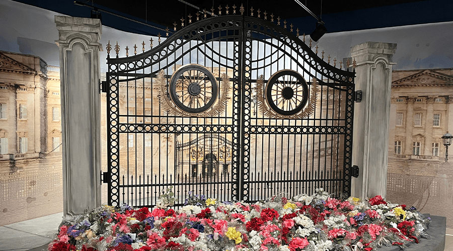 A recreation of the black Buckingham Palace gates after Princess Diana died with pink, blue, yellow and white flowers displayed in front of it and a screen picture of Buckingham Palace in the background.