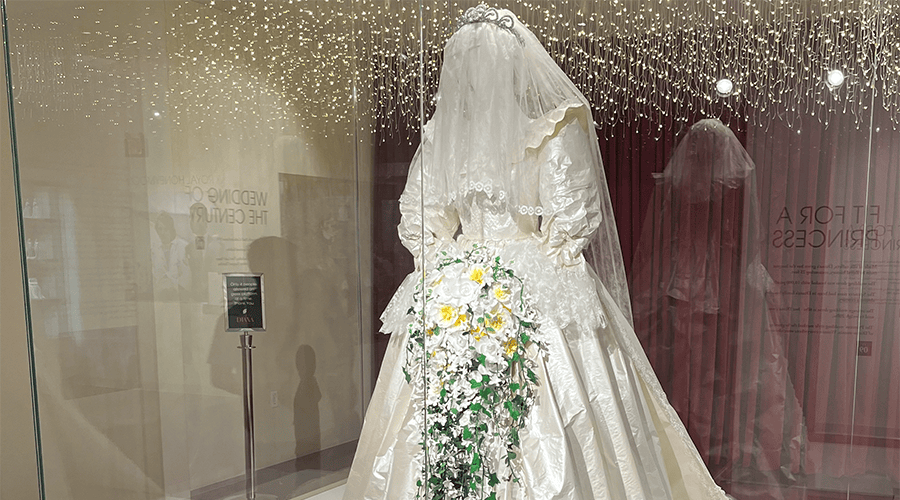 A recreation of Princess Diana's wedding dress in a glass case on display at the Princess Diana & The Royals Exhibition in Las Vegas  with a white and yellow floral arrangement.