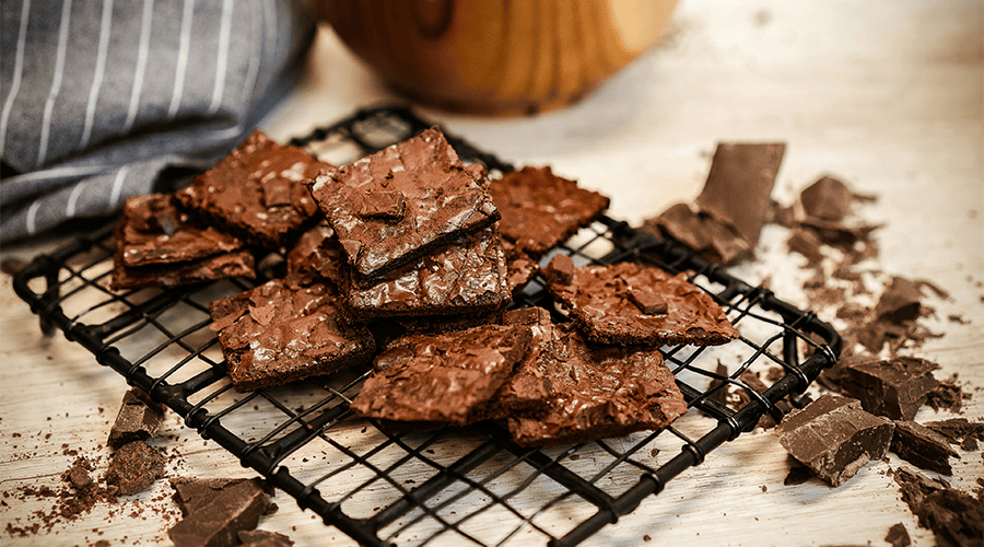 A batch of chocolate brownies on top of a black cooling grid with pieces of broken chocolate surrounding the cooling grid and a grey cloth with white lines in the background.