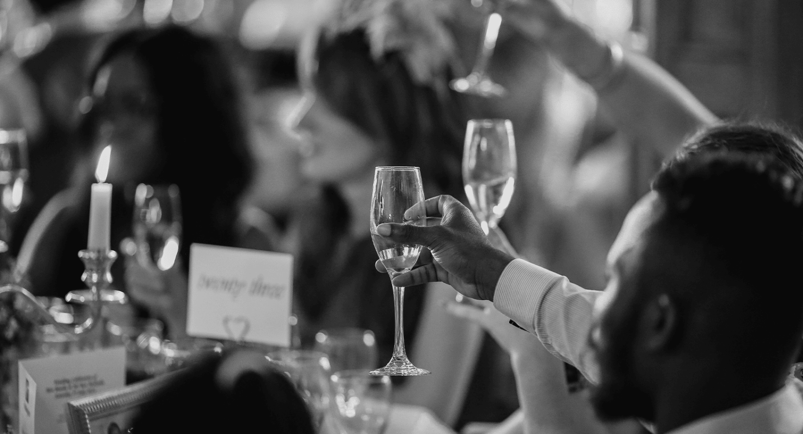 A black and white image of a male and female wedding party holding champagne glasses in the air for a toast with candles and a table name holder on the table.