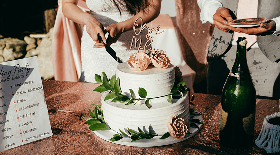 A bride and groom cutting their two-tier wedding cake with green flower decorations, a Mr & Mrs cake topper, and a bottle of champagne with an order of service on the left.