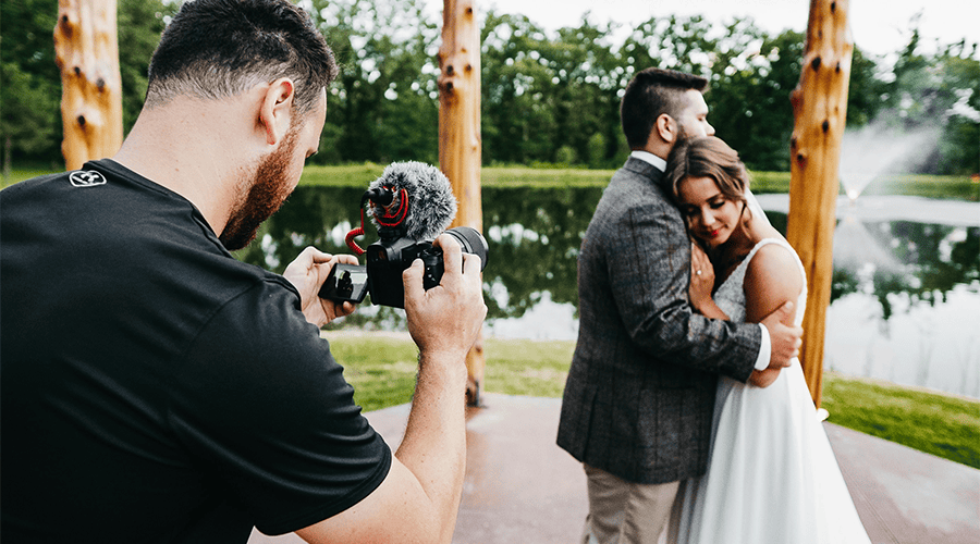 A videographer holding a black video camera wearing a black t-shirt filming a bride and groom next to a lake and green trees in the background.