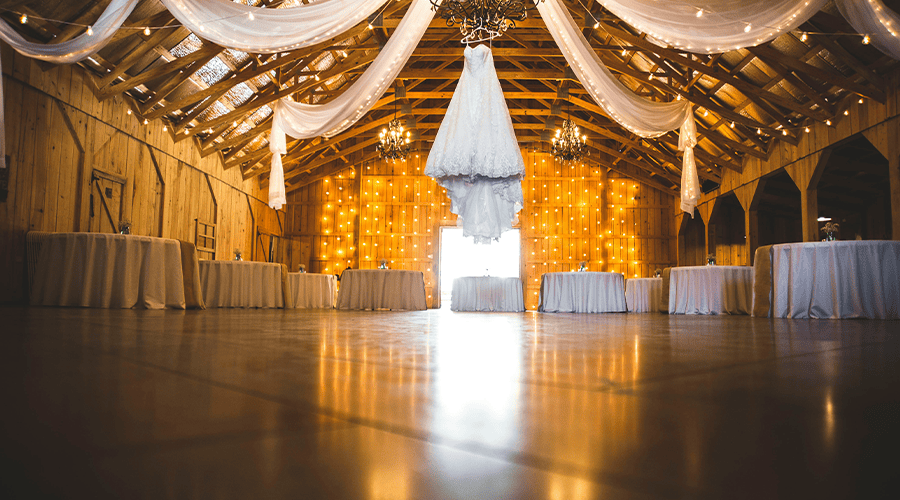 An empty wedding venue with white linen tables around the edge of the floor and white linen decorations and lights hanging from the ceiling.