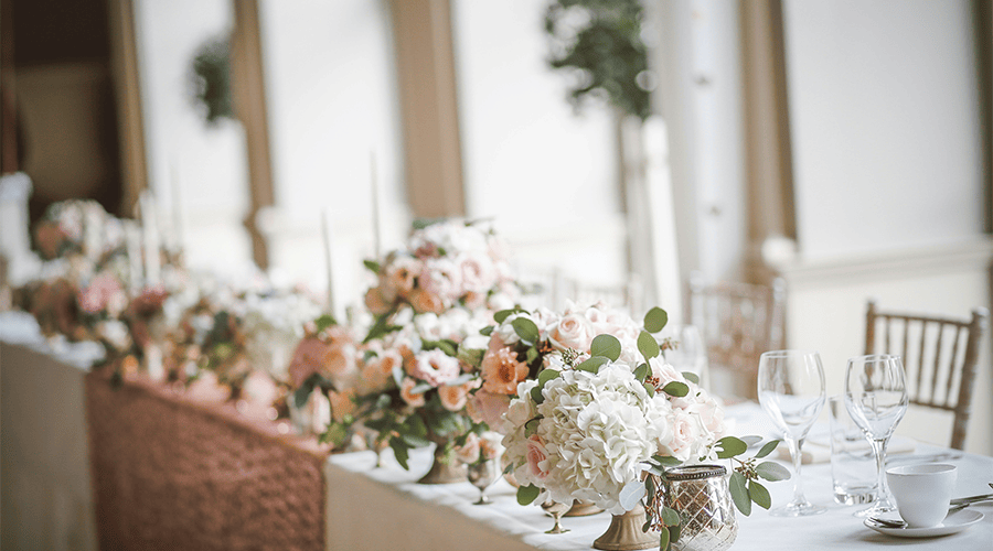The head table at a wedding with light pink, cream and white flower arrangements, wine glasses, cutlery and chairs against a window in the background. 