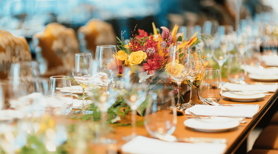 A long wedding breakfast table with a yellow and red flower arrangement in the middle, wine glasses, white plates and cutlery as well as chairs in the background.