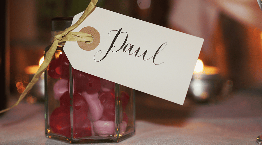A wedding favour jar of red and white sweets in a glass jar with a gold ribbon and name tag with "Paul" on it on top of a white linen tablecloth. 