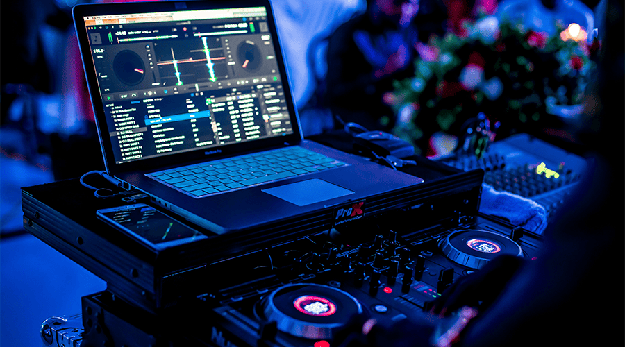 A laptop and phone on top od a DJ deck at a wedding with bright lights and buttons and guests dancing in the background.