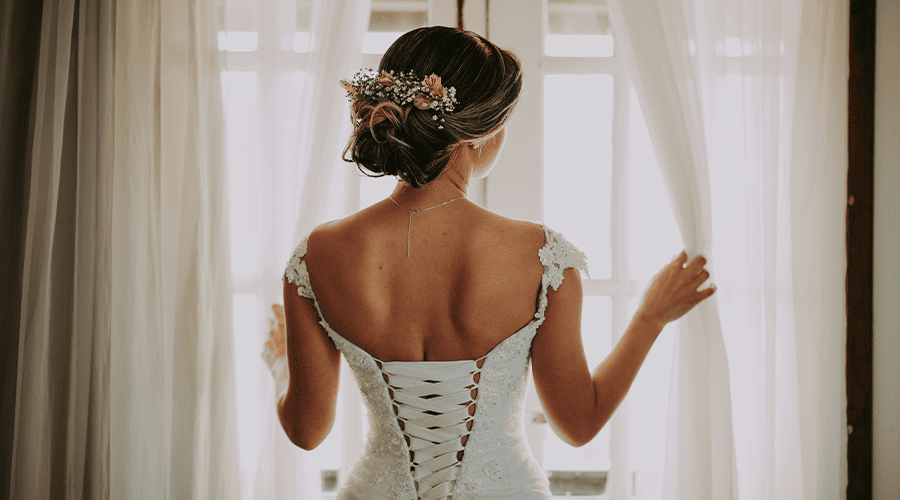 A bride wearing a white bridal gown with a strappy back fastening and a flower hair arrangement opening the white linen nets looking out of the window. 