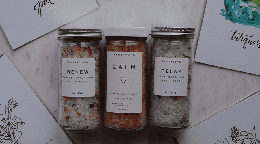 A selection of three bath salts for wedding hampers in plastic containers against a marble effect background and white cards with green and black and white drawings. 