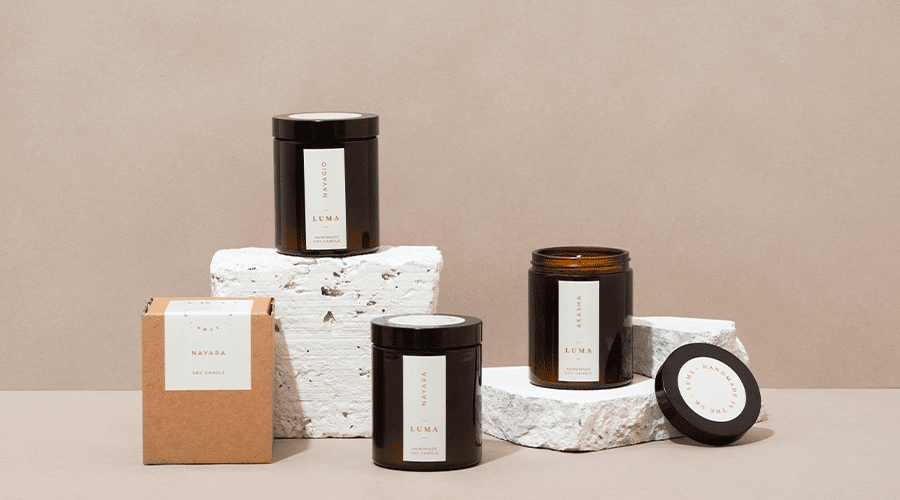 Four jar candles in front of a beige background and on top of a beige counter with one candle in a box, all candles with white labelling and two candles on top of white platforms.