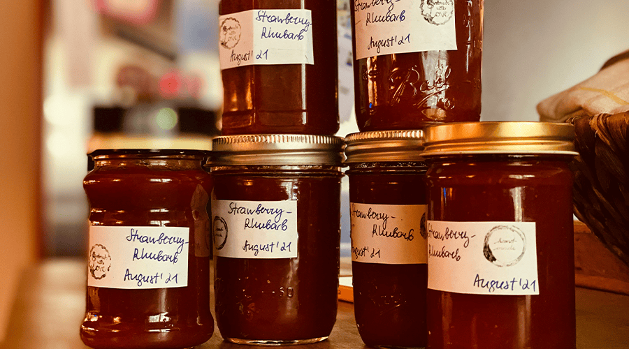 Five jars of strawberry and rhubarb jam with stickers on the front of each one and gold lids against a blurred background.