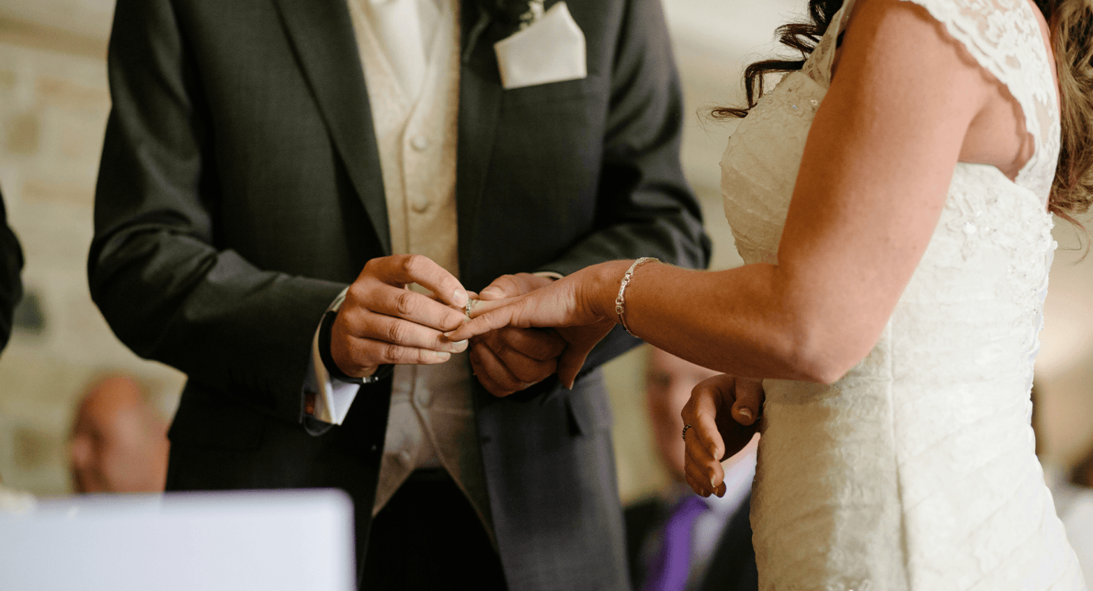A groom holding the bride's hand at the altar and placing a ring on her ring fingers with the froom wearing a black suit, white shirt and pocket square.