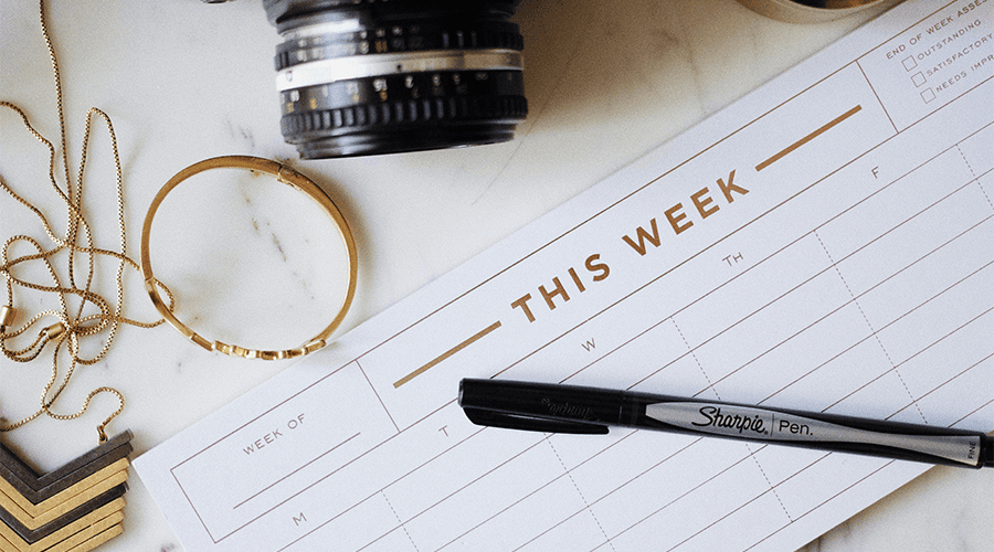 A weekly planner with gold lettering on top of a marble effect counter with a Sharpie pen, black camera and gold-coloured jewellery. 