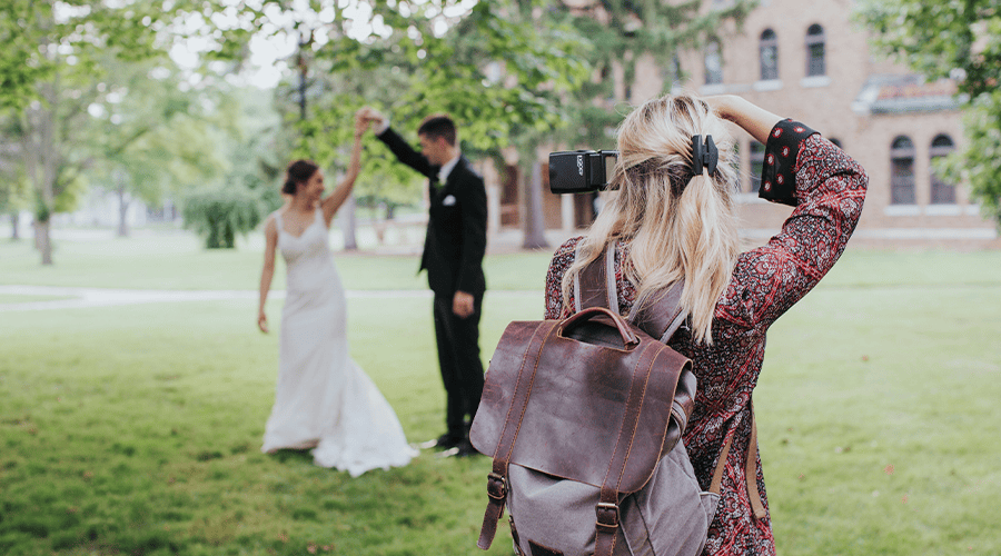 A female photographer wearing a brown backpack and printed long sleeve top holding a camera and photographing a couple on their wedding day on a green lawn against a backdrop of trees and a building.