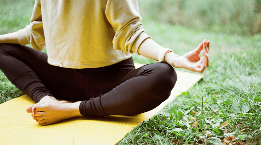 A female relaxing on her wedding day wearing a rolled-up sleeved top and dark leggings posing on a yoga mat on a green lawn with her hands in a yoga position. 