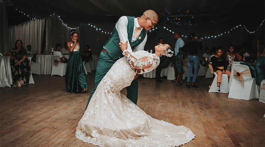 A bride and groom at their first dance on their wedding day with the bridge wearing a white gown and groom wearing a emerald green suit with guests around the dance floor sitting at tables.
