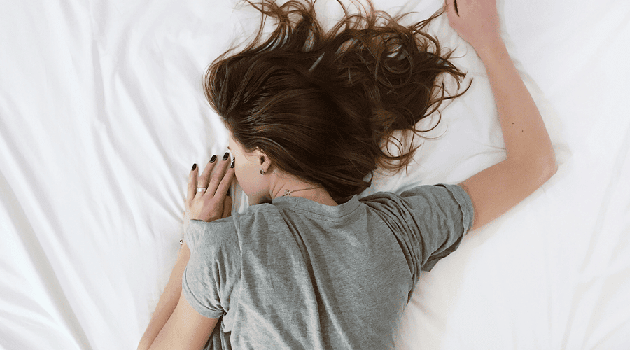 A female with brunette hair wearing a grey t-shirt and wedding ring sleeping on white sheets face down with her right arm up and left arm by her face.