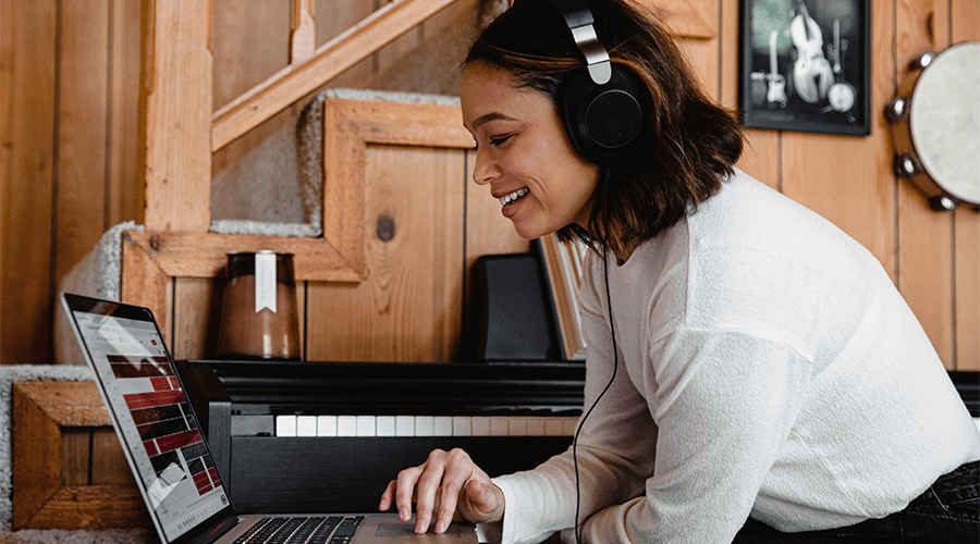 A female with brunette hair wearing a long sleeve white top and headphones and using her MacBook laptop listen to a podcast before her wedding day against a wooden staircase.