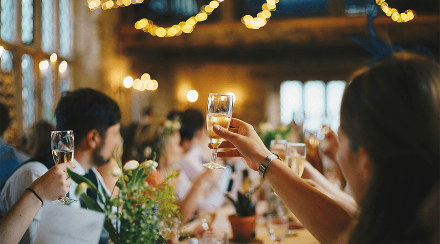 Male and female guests at a wedding venue holding glasses of champagne in the air for a toast with floral decorations and lights hanging from the ceiling.