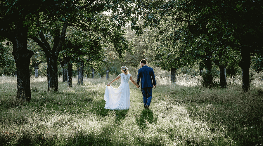 A bride and groom walking hand-in-hand against green trees and long green grass with the bride holding the bottom of her white wedding dress to the left side.