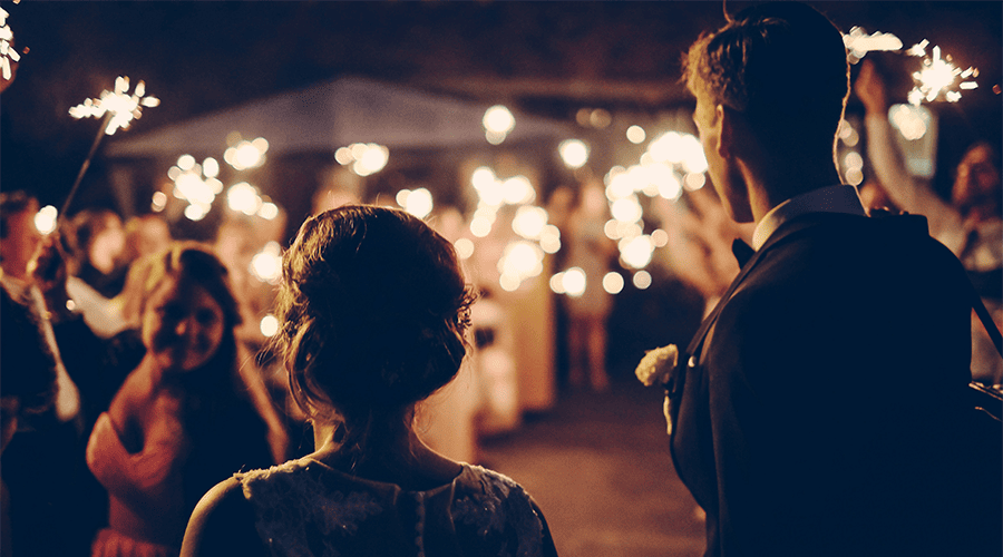 A bride and groom walking into their wedding venue with guests holding sparklets and the groom wearing a button hole and bridge with her hair up. 