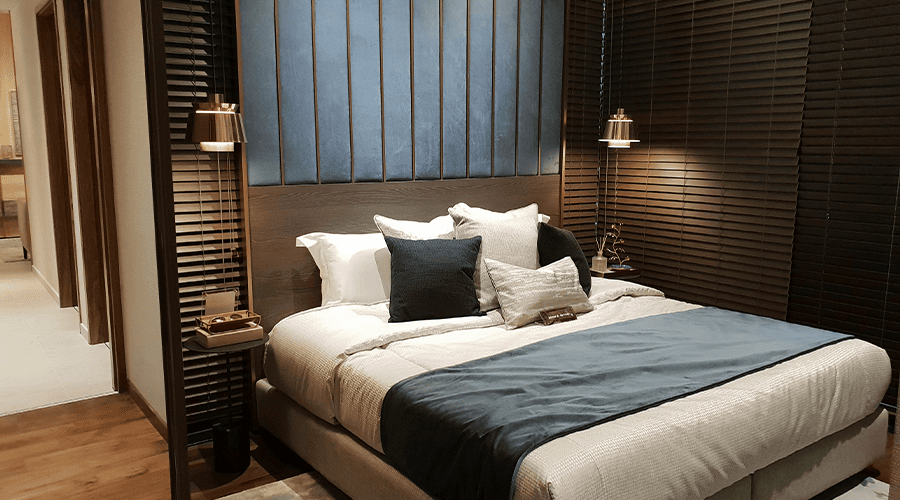 A bed at a wedding venue accommodations with blue and grey/blue pillows with a dark headboard and lights on either side of the bed and a wooden floor.