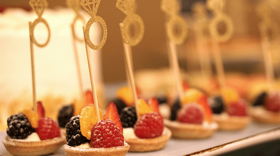 Strawberry and blackberry tartlets with a gold wedding ring decoration on a stick at the top of each tartlet at a wedding venue.