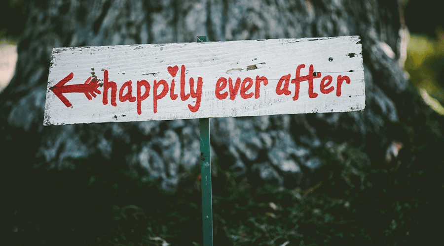 A "happily ever after" sign at a wedding venue with red font on a white background with a arrow pointing to the left against a backdrop of a tree trunk.