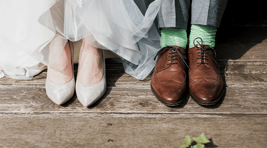 A bride and groom's shoes with the bride wearing off-white heels and white wedding dress and the groom wearing brown brogues and green socks on a wooden floor.